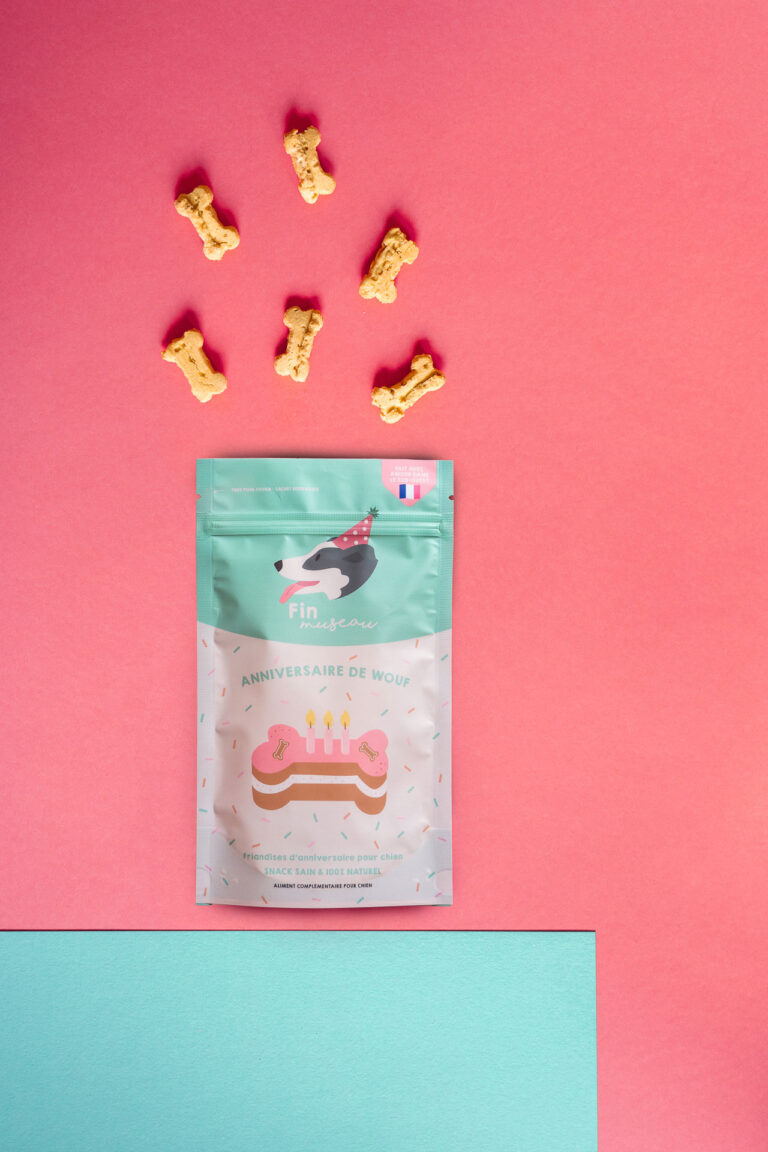 Cette image, magnifiquement capturée par un photographe professionnel spécialisé dans les mariages et familles des Landes, met en scène un paquet de friandises pour chiens intitulé "Anniversaire de Wouf", illustré d'un chien et d'un gâteau, tandis que des friandises en forme d'os sont artistiquement dispersées sur un fond rose, évoquant une atmosphère douce et festive parfaite pour une séance photo unique dans cette belle région.