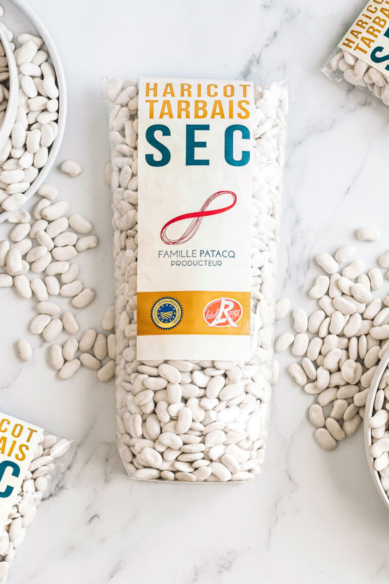Sur une élégante surface en marbre, un paquet de haricots blancs Tarbais séchés est délicatement disposé, avec quelques haricots éparpillés et des assiettes partiellement visibles, illustrant le talent artistique et l'œil raffiné d'un photographe professionnel spécialisé dans les mariages et les séances de famille dans les Landes.