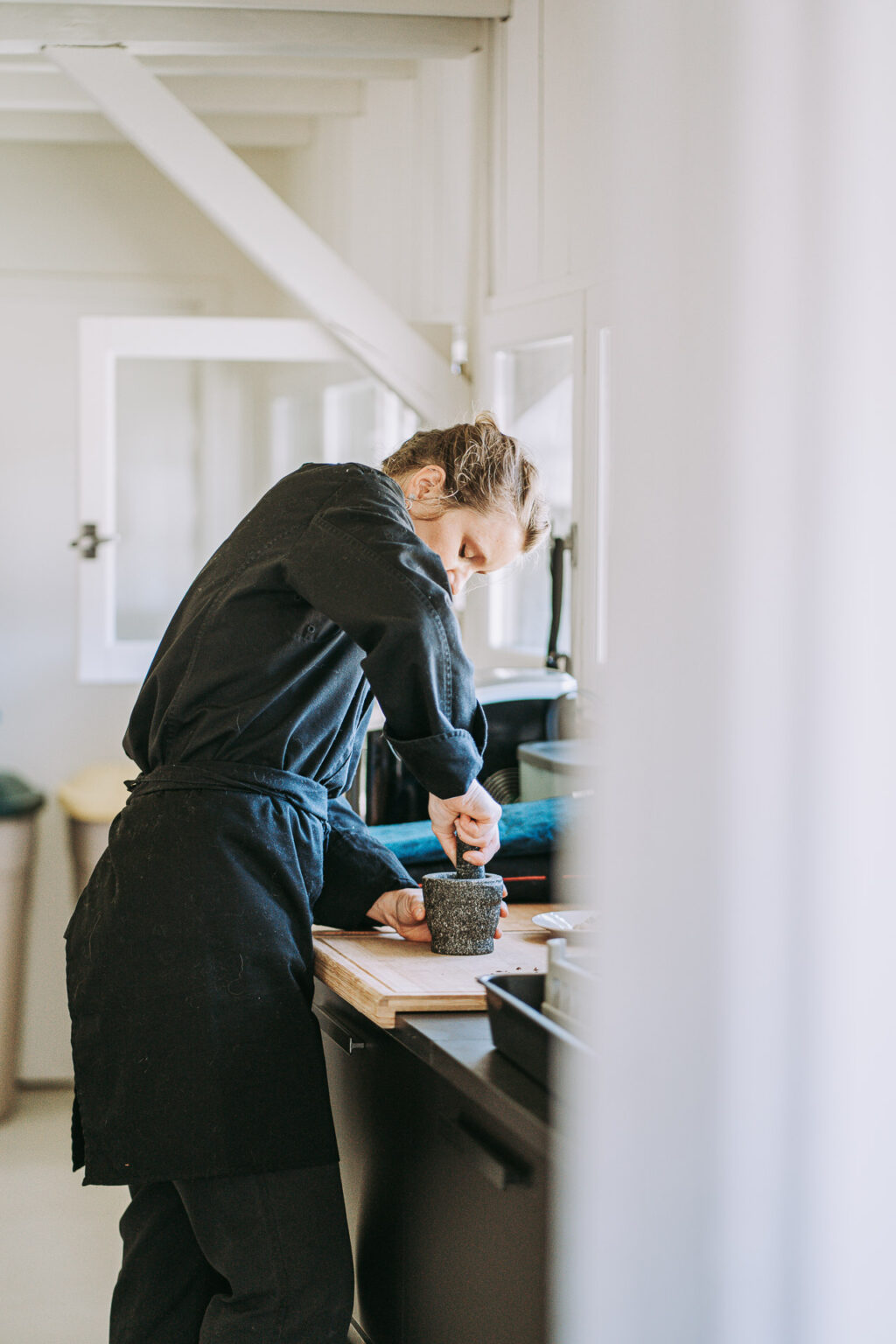 Sur un comptoir de cuisine baigné par la lumière du soleil, une personne portant un tablier noir utilise habilement un mortier et un pilon, illustrant une scène intime et authentique qui pourrait être magnifiquement immortalisée par un photographe professionnel spécialisé dans les mariages ou les portraits de famille dans les Landes, où l'art de capturer des moments précieux en toute simplicité est pris.