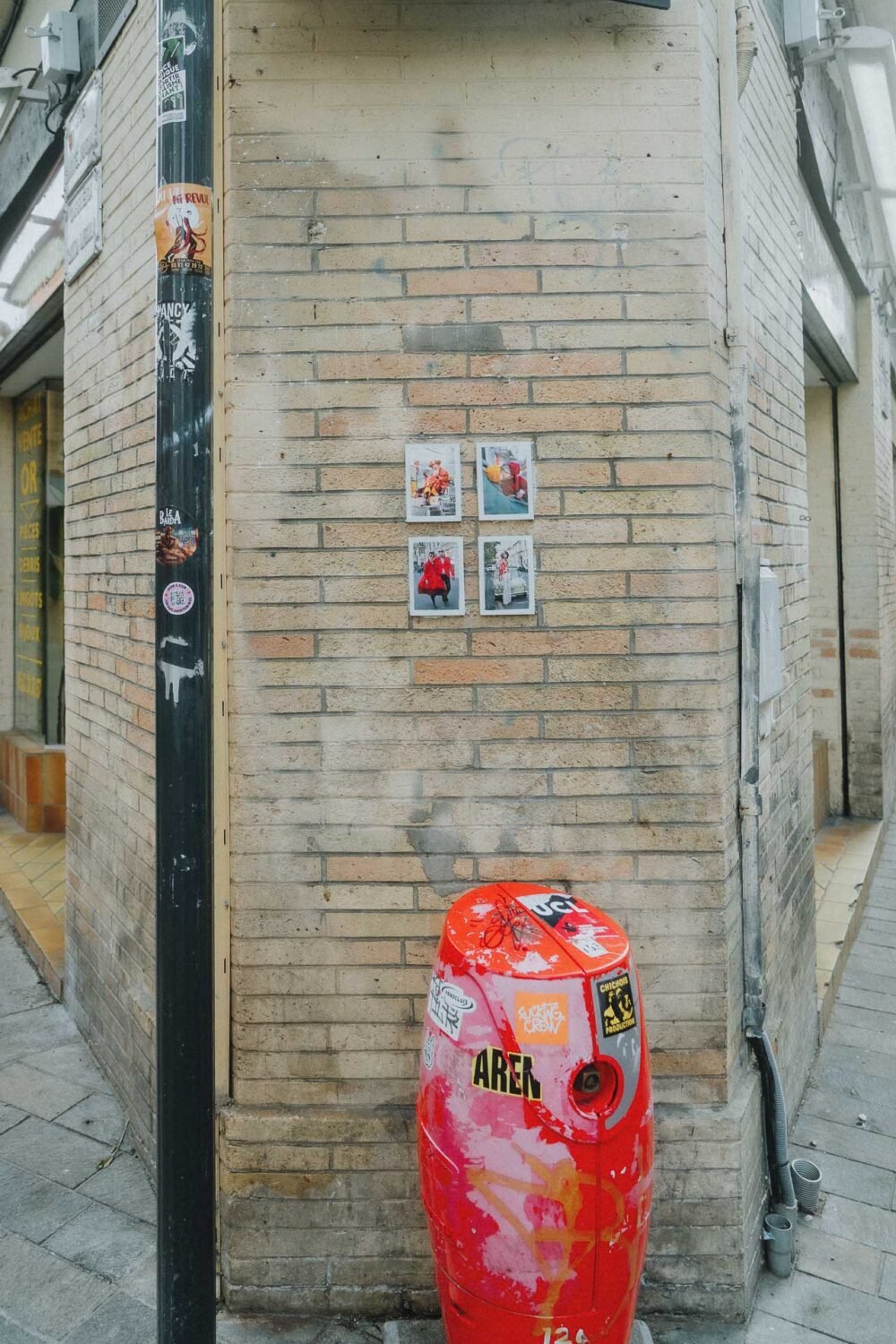 Cette scène urbaine, capturée par un photographe de mariage ou de famille dans les Landes, met en valeur un poteau rouge vif orné d'une multitude d'autocollants, se dressant fièrement près d'un mur de briques affichant quatre petites affiches ; à proximité sur le trottoir citadin de Dax dans les Landes, un autre poteau noir expose également une collection éclectique d'autocollants, créant ainsi une toile urbaine dynamique et colorée qui rappelle le style vibrant propre à Helidax.