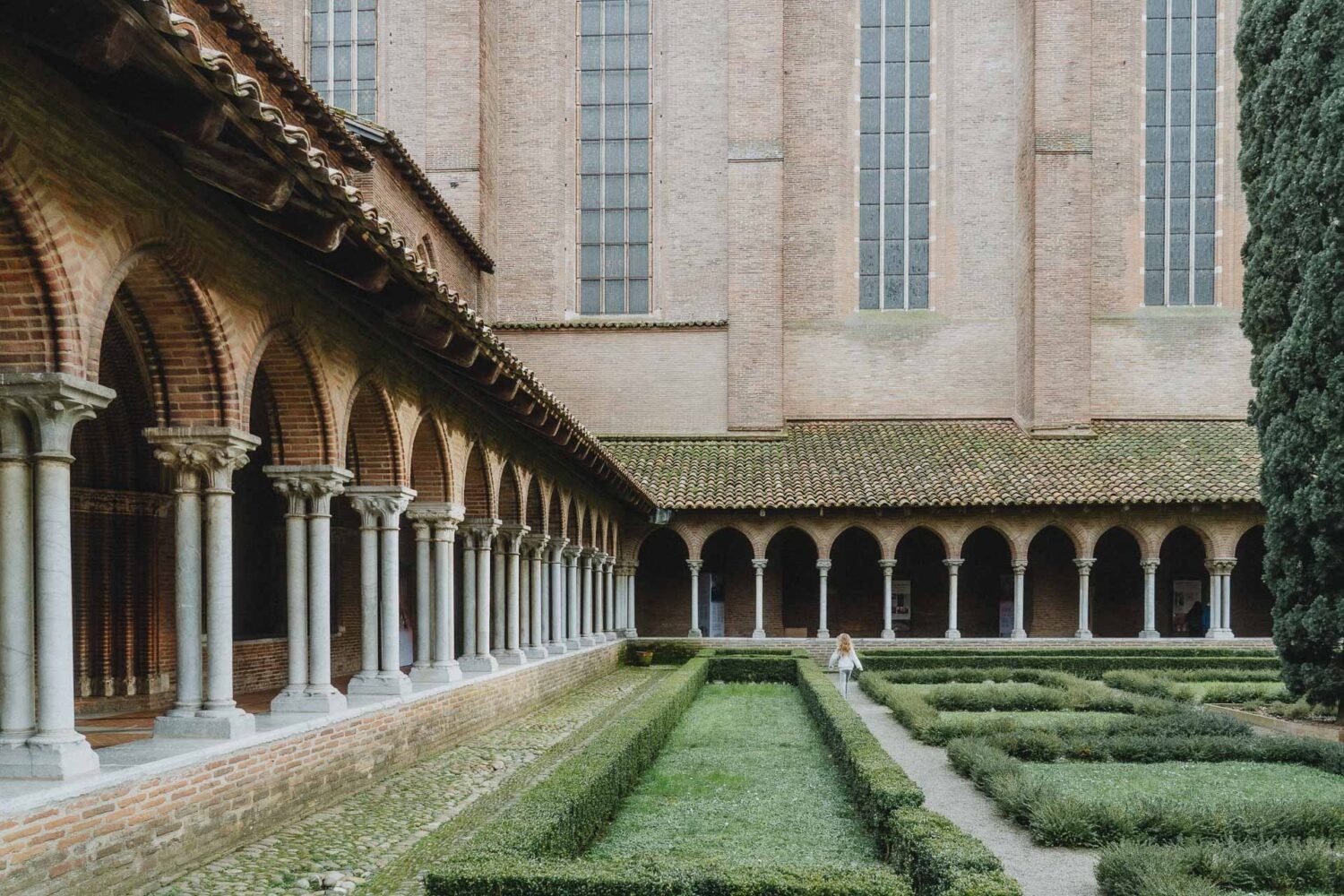 Dans les Landes, un photographe de mariage ou de famille capturerait parfaitement l'instant où une personne se promène avec élégance dans une cour bordée de colonnes en arcades et de haies soigneusement taillées, adjacente à un grand bâtiment en briques aux hautes fenêtres, tandis que le doux bourdonnement d'un hélicoptère Helidax résonne subtilement dans le ciel landais.