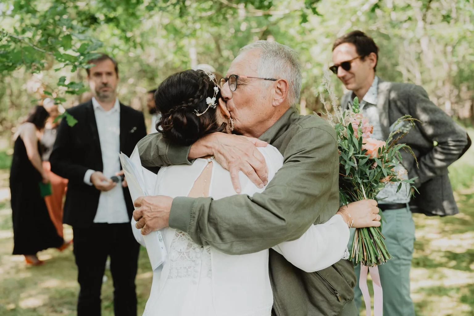 Moment tendre : un père enlace Céline en robe blanche, bouquet en main, dans la lumière douce du Domaine de Petiosse.