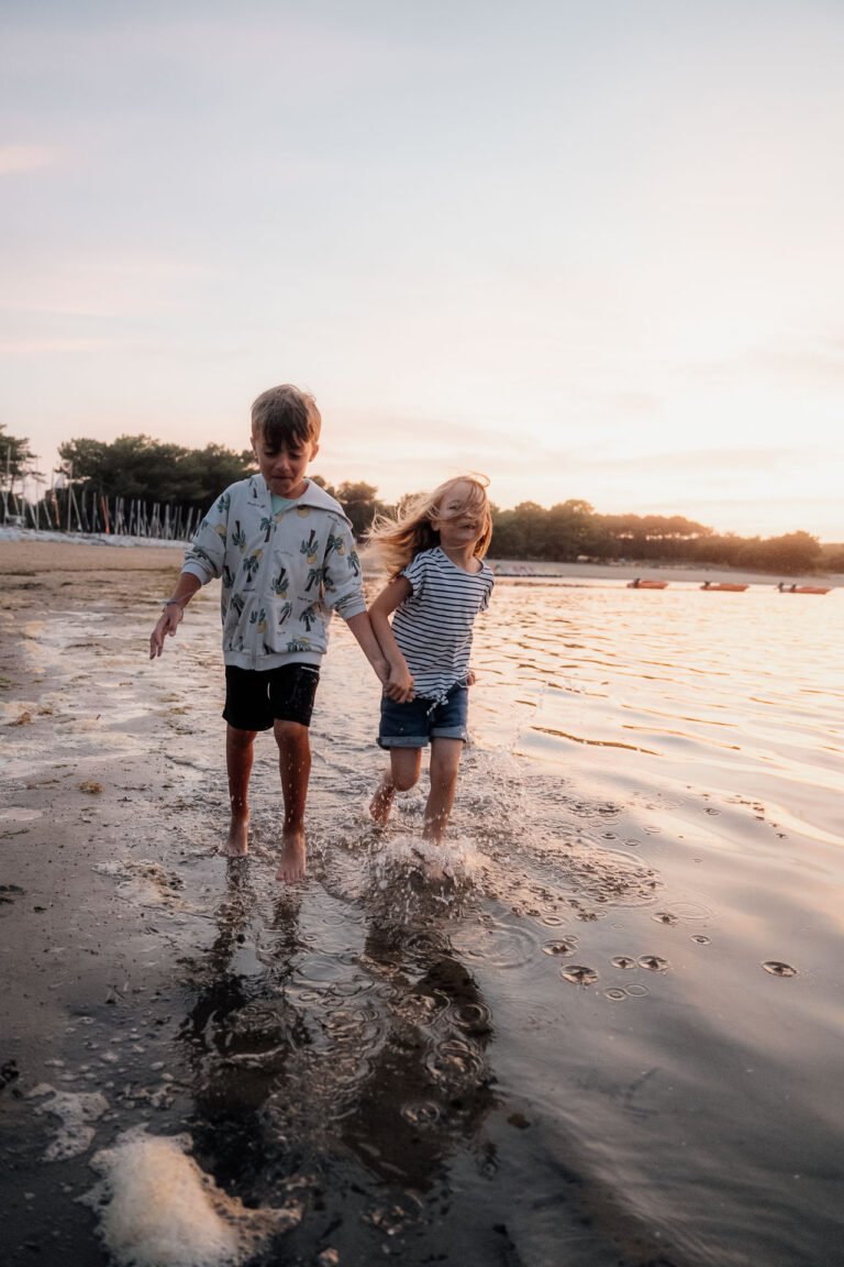 Deux enfants main dans la main traversent l'eau au coucher du soleil, instant doux pour une photo famille authentique dans les Landes.