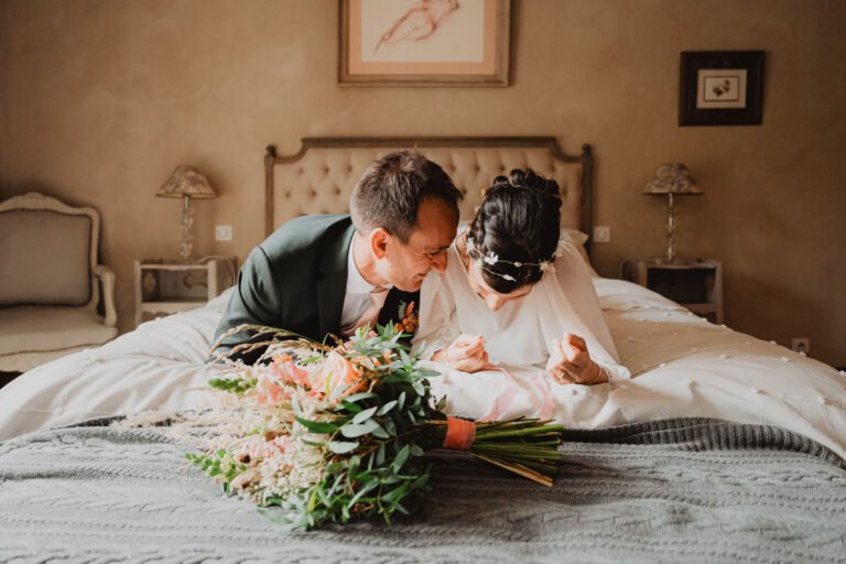 Céline et Nicolas, tendrement enlacés sur un lit, bouquet devant eux, lumière douce du Domaine de Petiosse, ambiance intime.