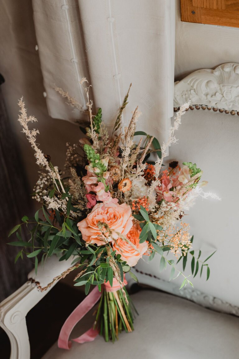 Délicat bouquet aux tons pêche et roses sur fauteuil raffiné, ambiance douce de mariage au Domaine de Petiosse dans les Landes.