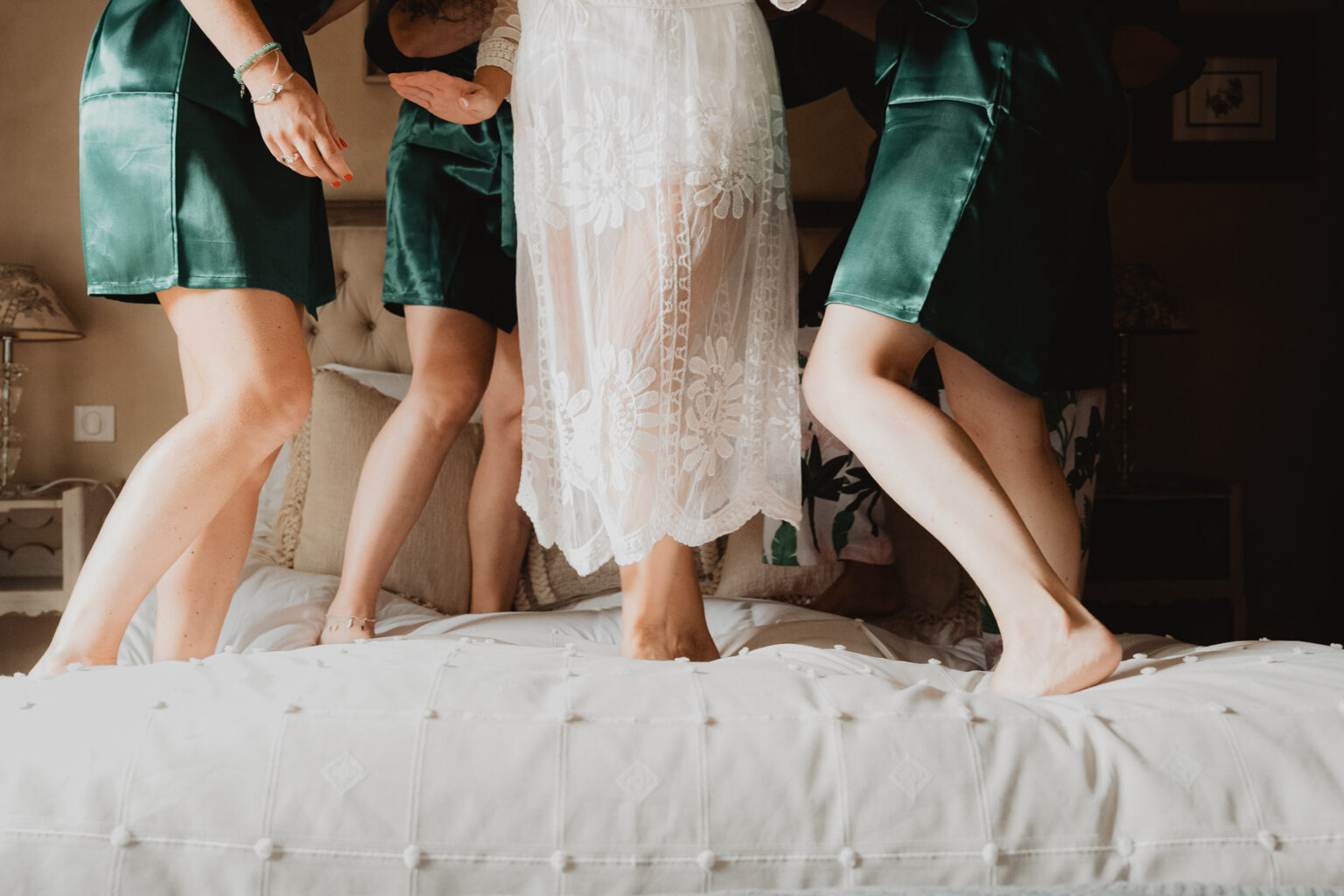 Céline en robe blanche entourée d'amies, pieds nus sur un lit, lumière douce, moment complice au domaine de Petiosse.
