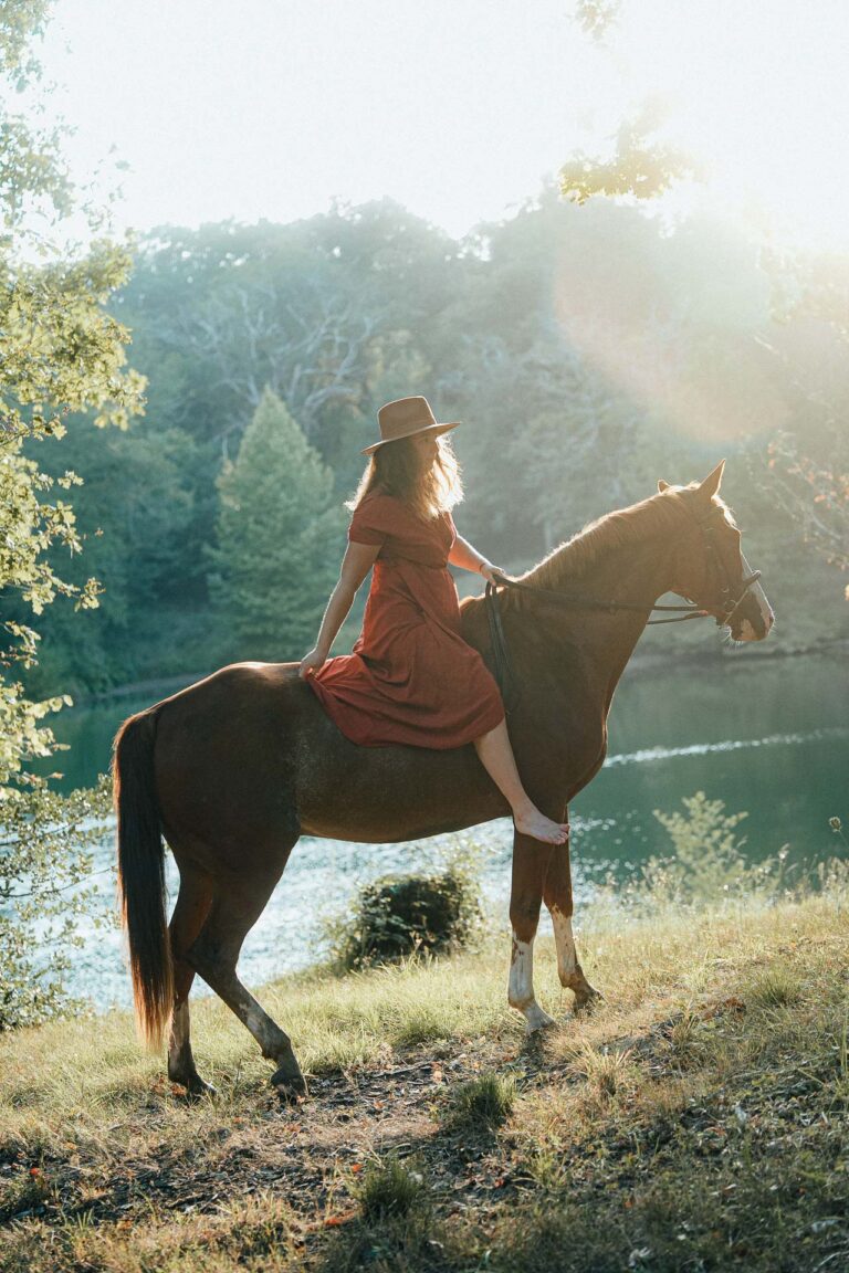 Moment tendre d'une femme en robe rouge sur un cheval, pieds nus, près d'un lac landais baigné de lumière pour mariage champêtre.