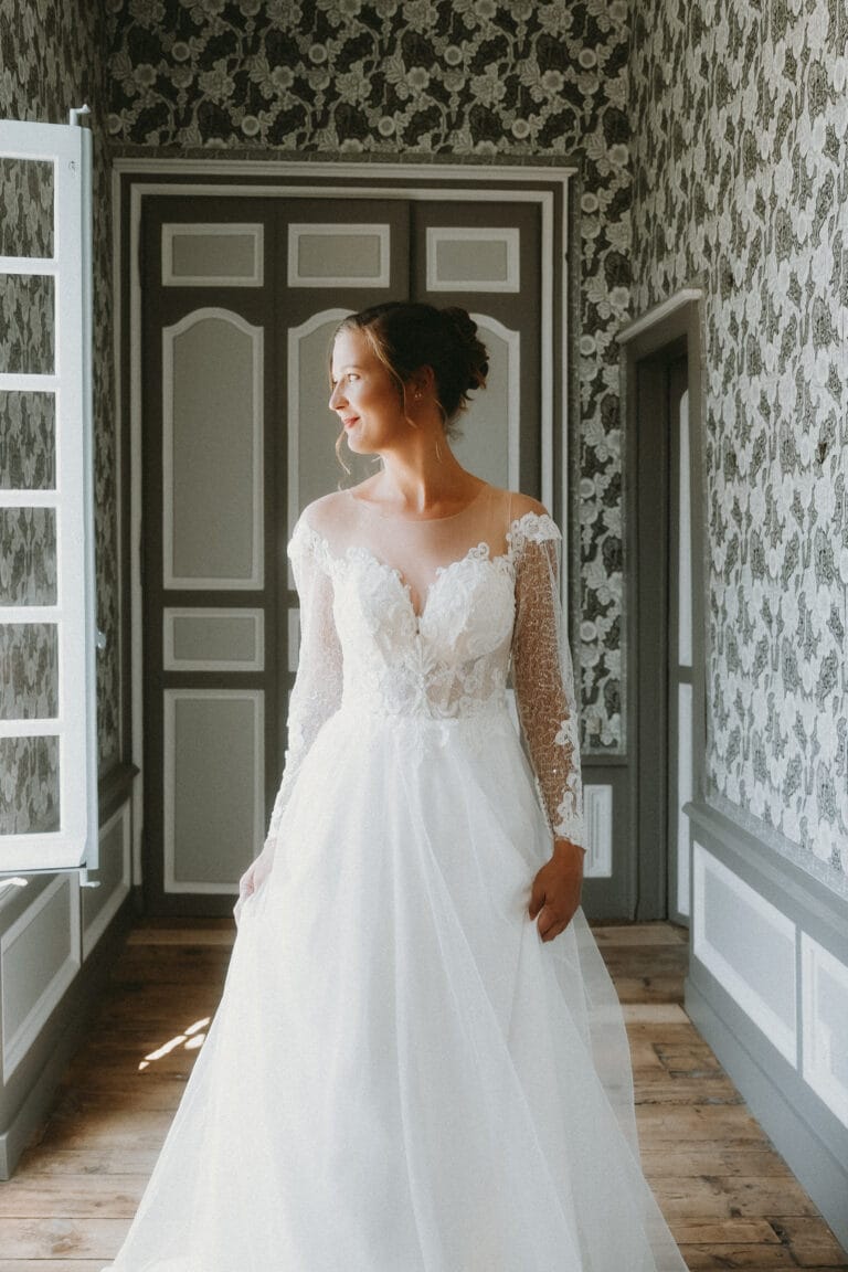 Vous êtes radieuse dans votre robe en dentelle, baignée de lumière douce à la fenêtre le jour de votre mariage à Dax.