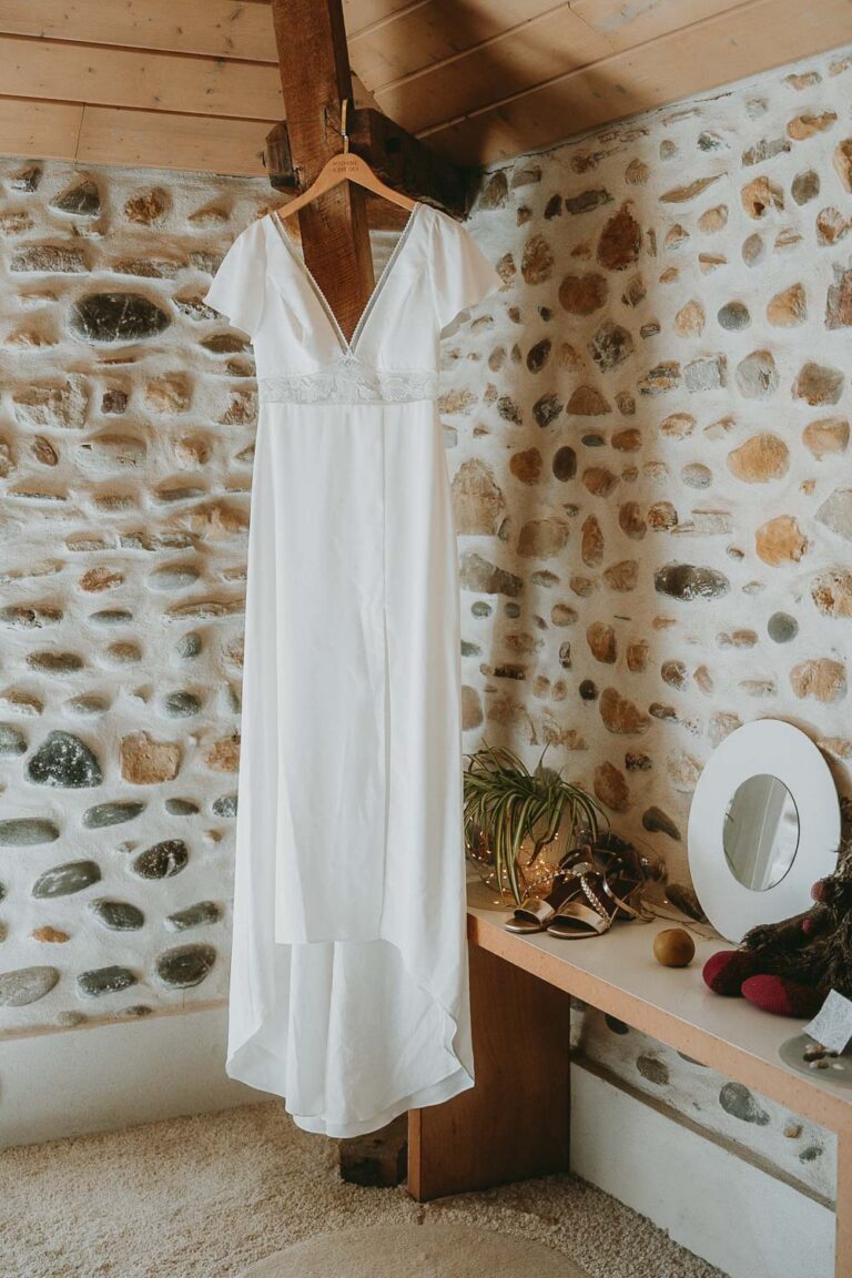Robe de mariée suspendue dans une pièce en pierre, ambiance douce et authentique des Landes, détails chaleureux de famille.