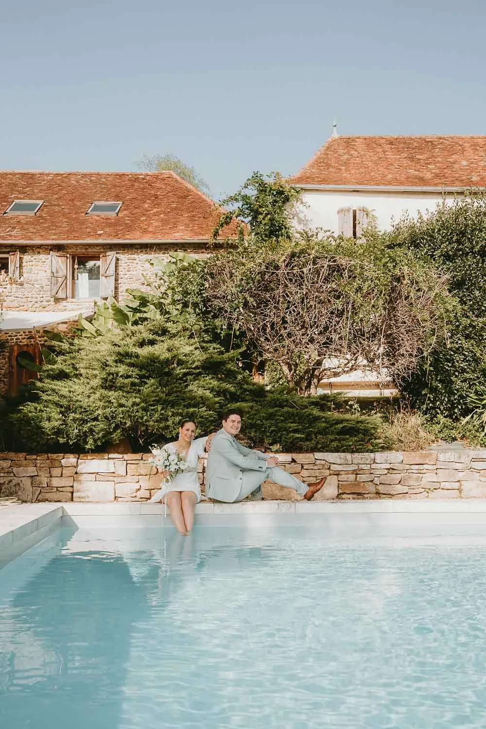 Instant tendre au bord d'une piscine, pieds dans l'eau, devant une bâtisse en pierre et verdure, ambiance Landes authentique.