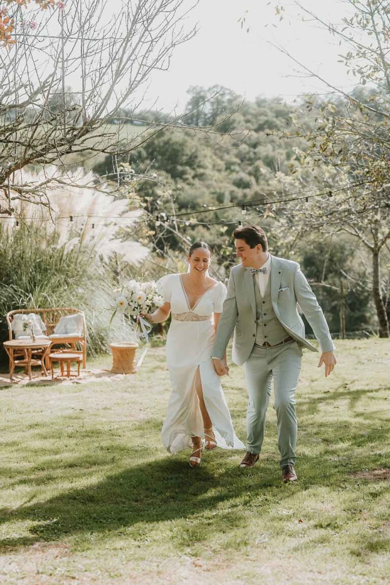 Un couple rayonnant marche main dans la main sous le soleil, entouré de verdure, capturé en douceur dans les Landes.