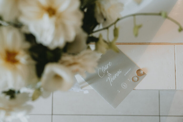 Carte de mariage posée sur carrelage ancien, alliances et fleurs blanches, ambiance douce et élégante des Landes.