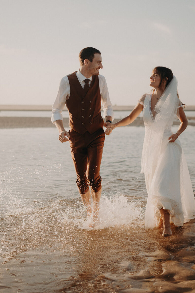 Un couple marche pieds nus dans l'eau au soleil couchant, main dans la main, instant complice capturé sur la plage des Landes.