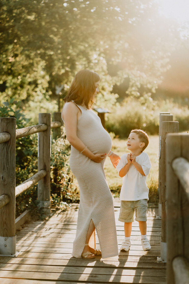 Maman enceinte en robe claire sur un pont en bois des Landes, regardant tendrement son fils sous la lumière douce du soleil.