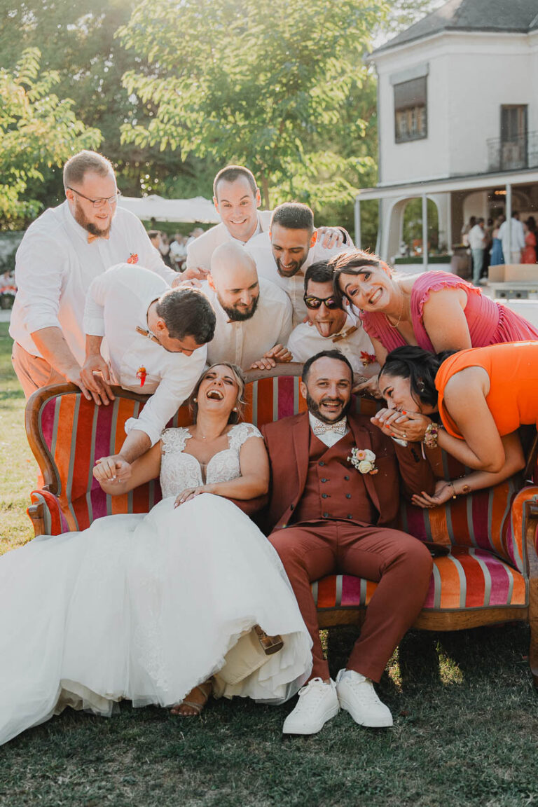 Photo de mariage dans les Landes : couple radieux entouré d'amis joyeux sur canapé rayé, ambiance douce et conviviale.