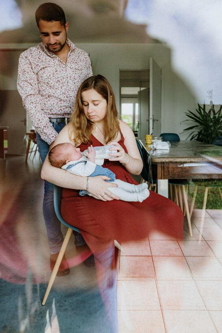 Tendre moment en famille, maman nourrit bébé au biberon, papa veille derrière. Ambiance chaleureuse dans une maison des Landes.