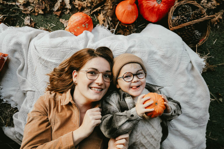 Instant tendre sur une couverture dans les Landes, mère et enfant souriants entourés de citrouilles, lumière douce d'automne.