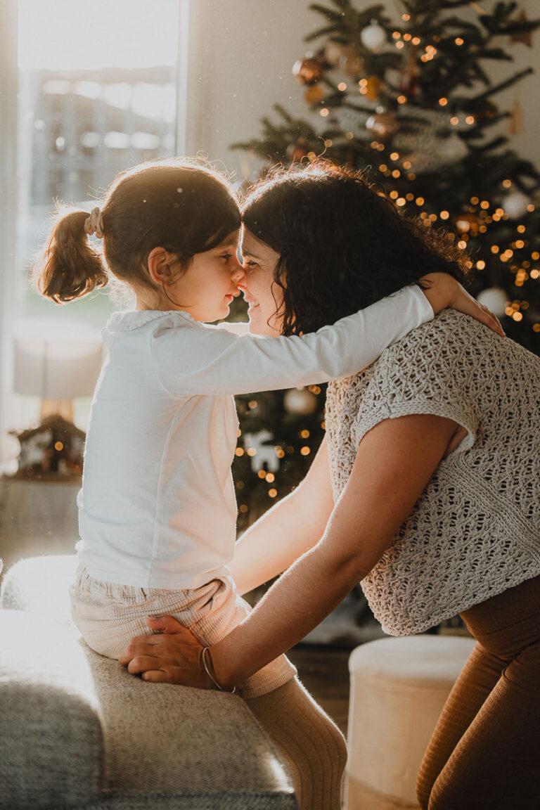 Douce connexion mère-fille devant le sapin, sourire complice et lumière naturelle, capturée par un photographe des Landes.