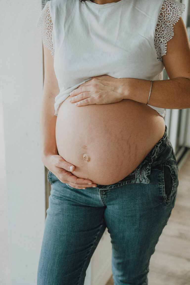 Une future maman en blanc et jean, caressant tendrement son ventre, pose chez vous à Dax pour un souvenir doux et authentique.