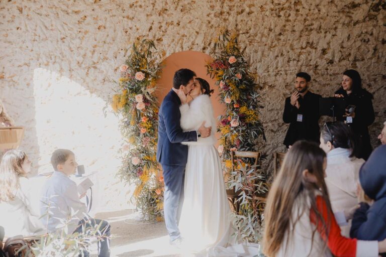 Un doux baiser sous une arche fleurie, entourés de proches dans une belle salle en pierre, souvenir intime des Landes.