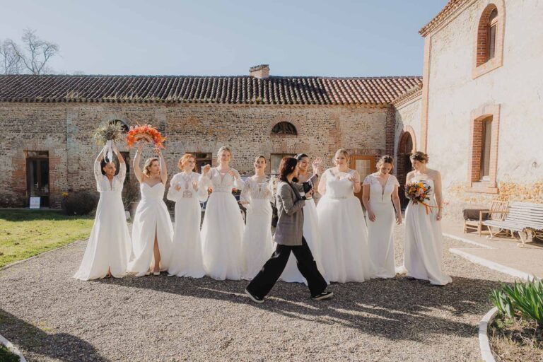 Huit mariées radieuses posent devant de vieux bâtiments sous le ciel bleu, une scène naturelle capturée dans les Landes.