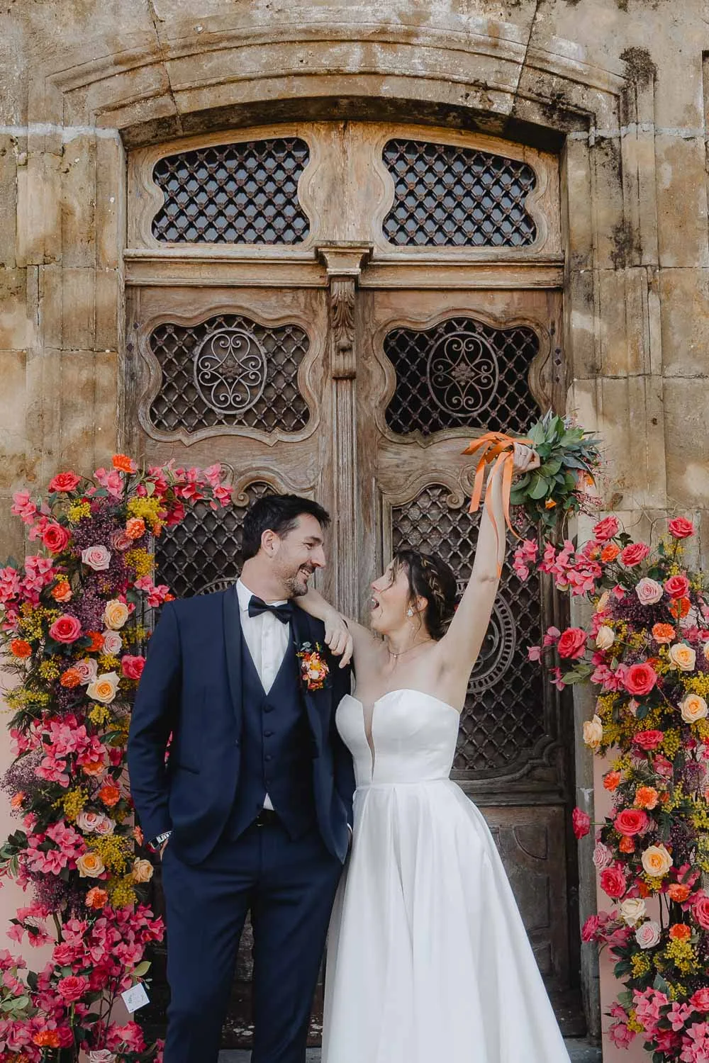 Un couple radieux devant une porte en bois, entouré de fleurs ; bonheur et complicité d'un mariage dans les Landes.