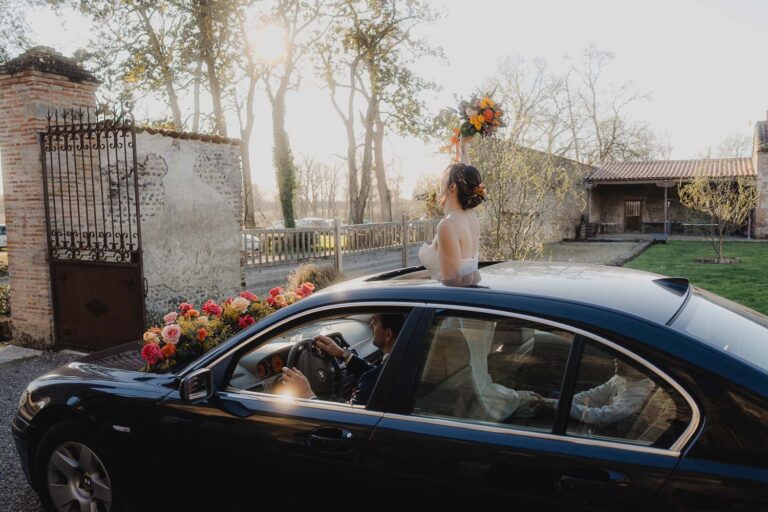 Une mariée rayonnante salue depuis une voiture fleurie, bouquet à la main, sous le soleil des Landes, ambiance douce et festive.
