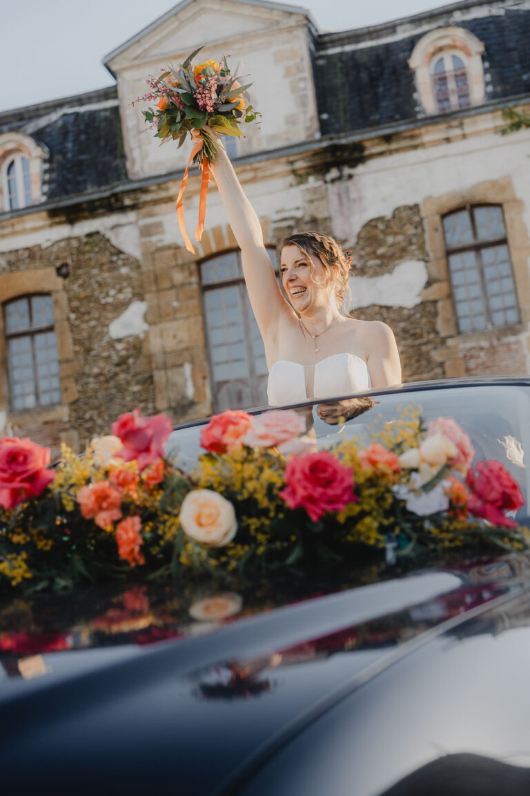 Une mariée radieuse en robe blanche sourit dans une voiture fleurie devant une bâtisse ancienne, immortalisée dans les Landes.