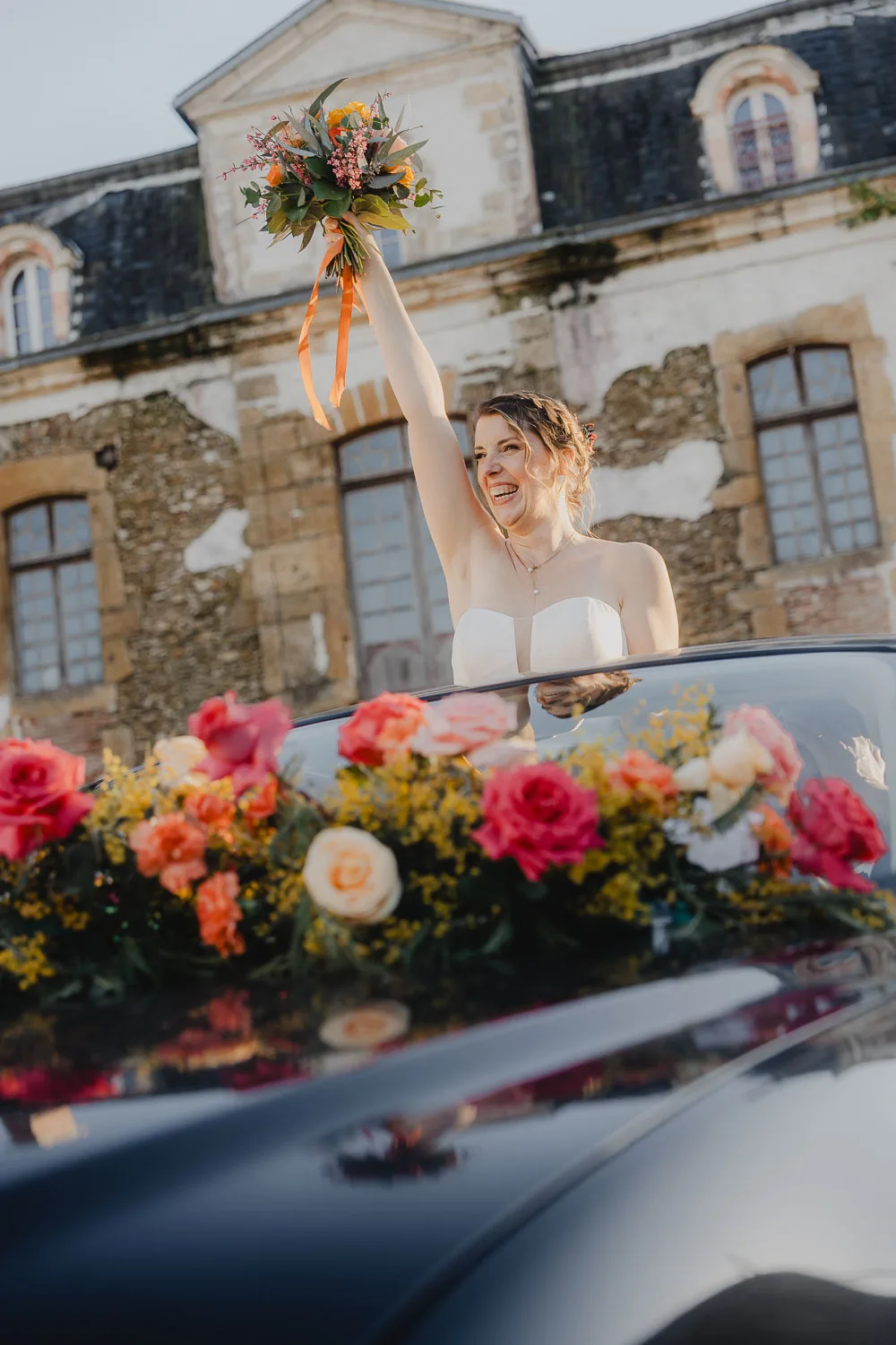 Le Jour J – Allison Micallef Photographe-782 Une mariée radieuse en robe blanche sourit dans une voiture fleurie devant une bâtisse ancienne, immortalisée dans les Landes.