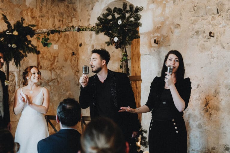 Un doux moment de mariage dans une salle en pierre, la mariée rayonnante écoute des chants, capturé dans l'ambiance landaise.