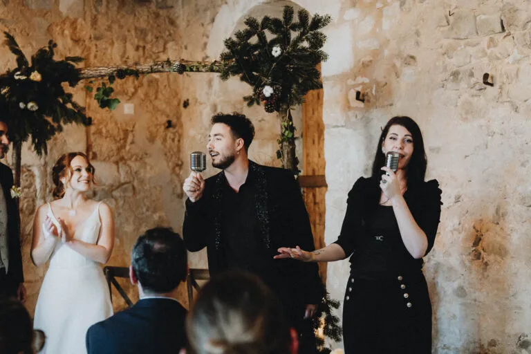 Un doux moment de mariage dans une salle en pierre, la mariée rayonnante écoute des chants, capturé dans l'ambiance landaise.