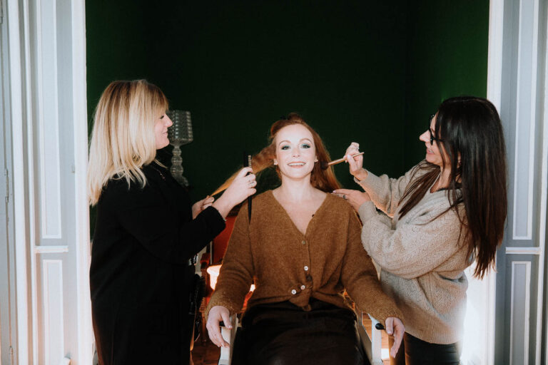 Dans une ambiance douce aux murs verts, une future mariée rayonne entourée de deux artistes beauté avant sa séance photo Landes.