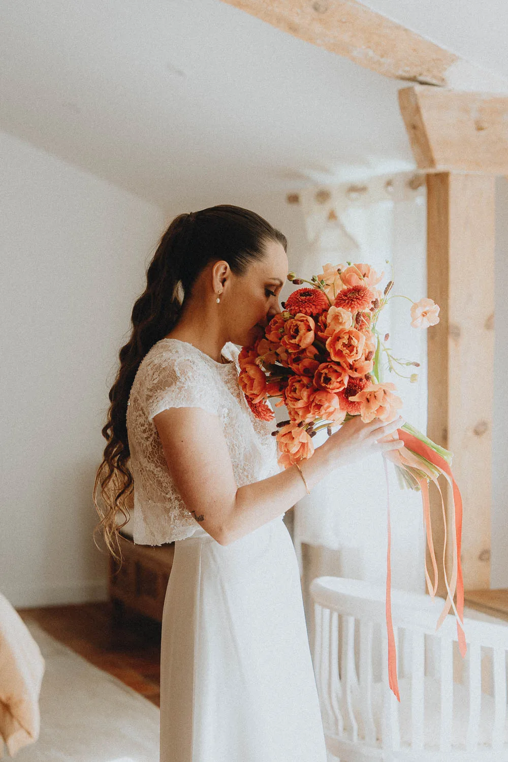 Mariée émue sentant son bouquet coloré sous poutres boisées, séance mariage authentique à Dax, Landes.