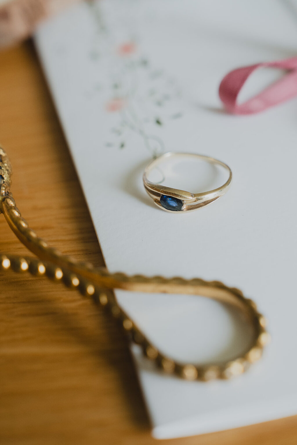 Bague dorée sertie d'une pierre bleue, posée sur une carte fleurie, un bijou doré et un ruban rose : une ambiance de mariage délicate à Dax.