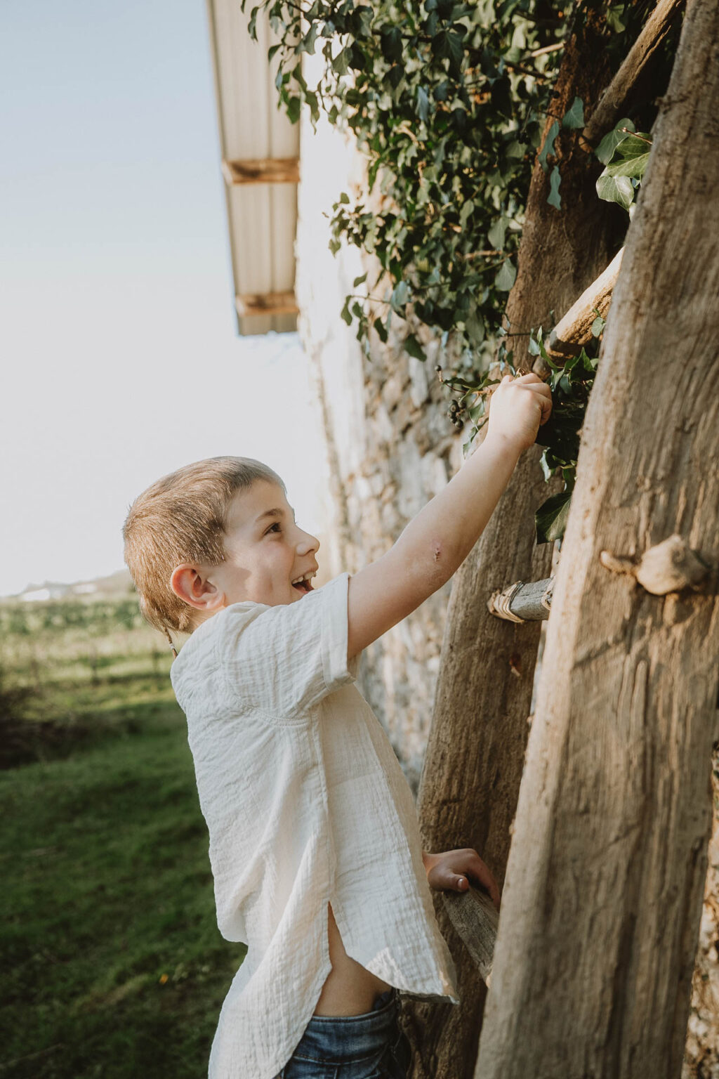 Un enfant s'amuse joyeusement sur une échelle en bois près d'un mur en pierre, séance photo en famille au naturel à Dax, dans les Landes.