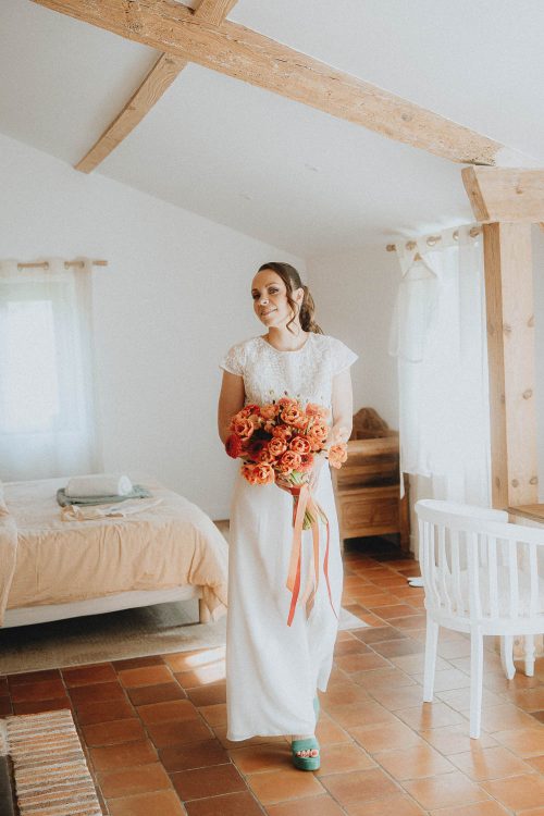 Sourire lumineux d'une future mariée aux Landes, bouquet orangé en main dans une chambre rustique pleine de douceur.
