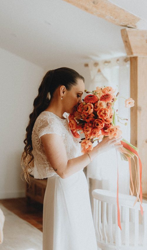 Mariée émue sentant son bouquet coloré sous poutres boisées, séance mariage authentique à Dax, Landes.