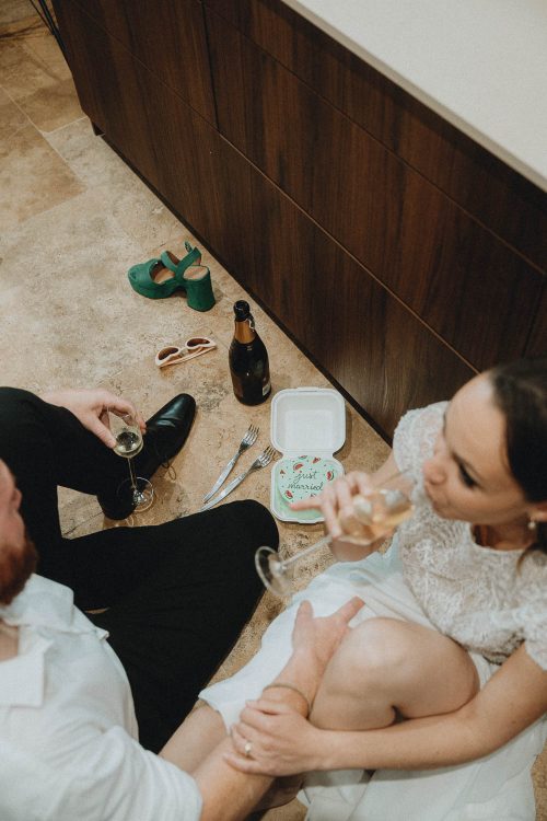 Jeune couple en tenue élégante, rient au sol de leur cuisine à Dax, champagne et gâteau "Just Married", chaussures posées nearby.