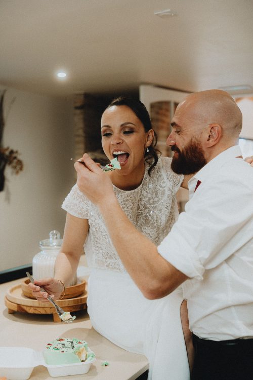 Moment complice lors d'un mariage à Dax : la mariée sourit, assise sur un comptoir, son époux lui offre du gâteau.
