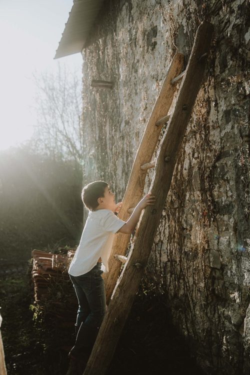 Un garçon grimpe à une échelle en bois appuyée contre un mur de pierre sous le soleil, séance photo en famille à Dax, dans les Landes.