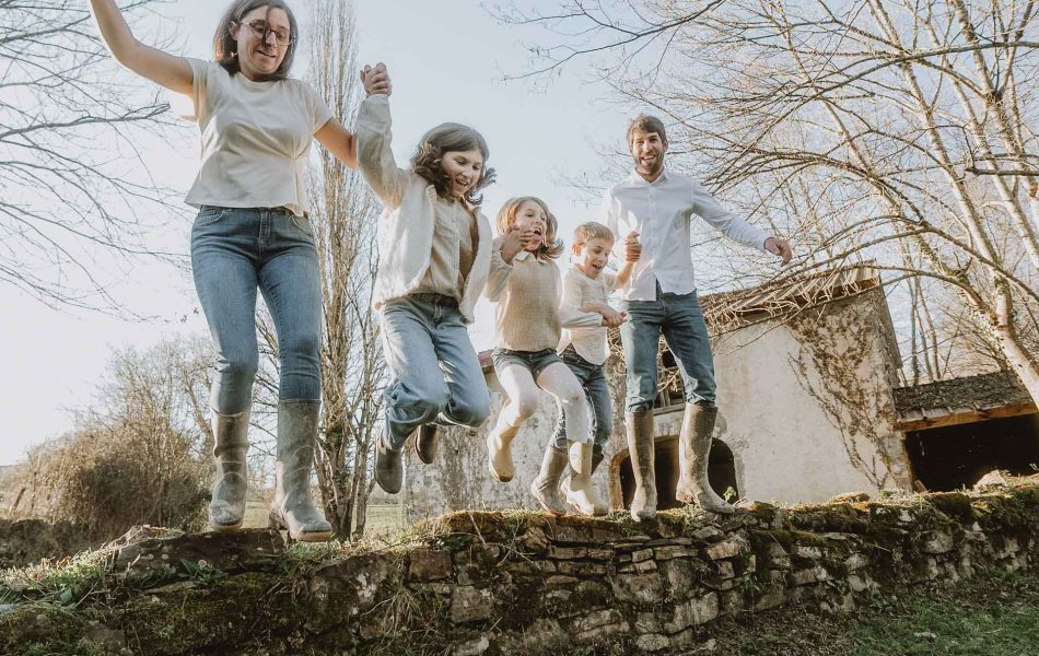 Une famille sautant main dans la main depuis un muret, éclats de rire au soleil lors d’une séance photo en famille près de Dax.
