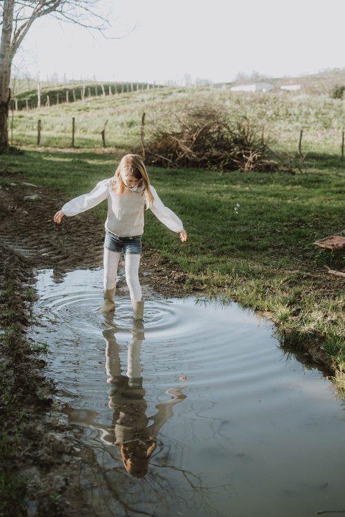 Une petite fille joue dans une flaque d'eau sur un sentier verdoyant, dans une ambiance joyeuse lors d'une séance photo en famille près de Dax, dans les Landes.