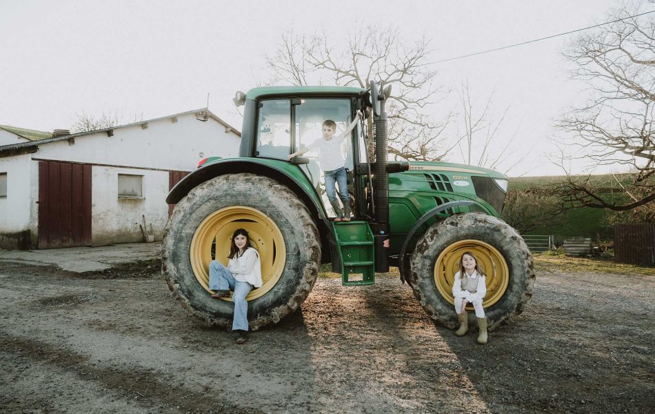 Une complicité immédiate lors d’une séance photo en famille à Dax : un trio joyeux autour d’un tracteur vert, près d’une grange et d’arbres.