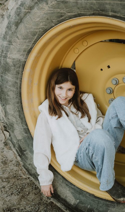 Petite fille souriante assise dans une roue de tracteur jaune, séance photo en famille pleine de joie à Dax, dans les Landes.