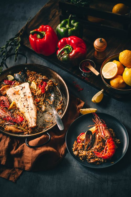 Cette image magnifiquement capturée par un photographe professionnel des Landes illustre une délicieuse paella de fruits de mer, incluant poisson et crevettes, artistiquement entourée de citrons, poivrons et épices colorées, avec une portion savamment dressée sur une assiette noire accompagnée d'une cuillère et d'une fourchette, reflétant le talent culinaire souvent souligné lors des mariages ou réunions familiales dans notre belle région landaise.