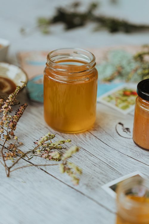 Imaginez ce pot de miel doré sur une table blanche, entouré de fleurs, ambiance douce et champêtre des Landes.