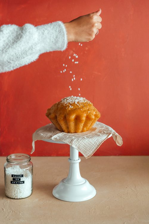 Une main saupoudre de sucre un gâteau doré sur présentoir, ambiance chaleureuse comme une fête de famille dans les Landes.
