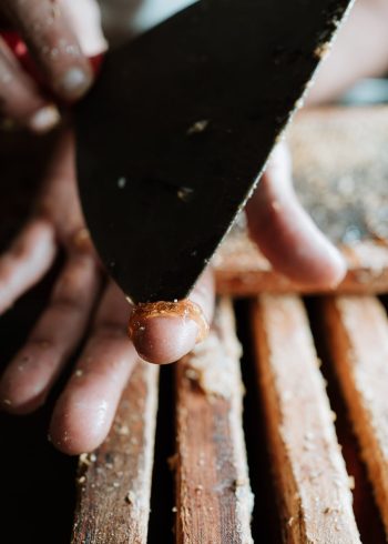 Dans les Landes, un photographe professionnel de mariage et de famille immortalise avec précision un gros plan captivant où des doigts habiles manipulent une planche en bois, étalant soigneusement de l'adhésif à l'aide d'un couteau à mastic, illustrant ainsi l'artisanat méticuleux qui peut ajouter une touche authentique aux photos de mariage et renforcer la beauté intemporelle des albums photographiques familiaux.